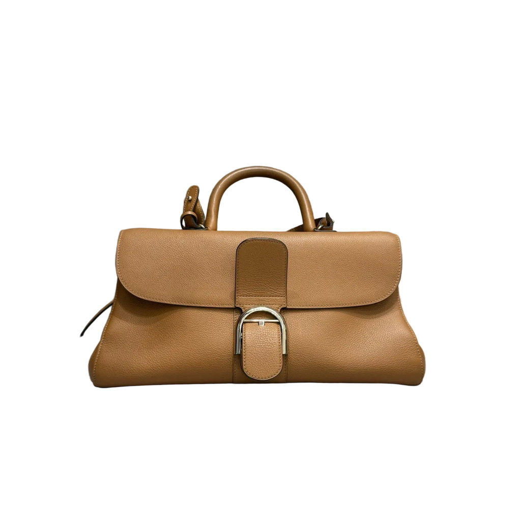 Front view of a Delvaux Brillant East/West handbag in tan box calf leather, featuring the iconic gold-tone D-buckle, a structured top handle, and a detachable shoulder strap on a white background.
