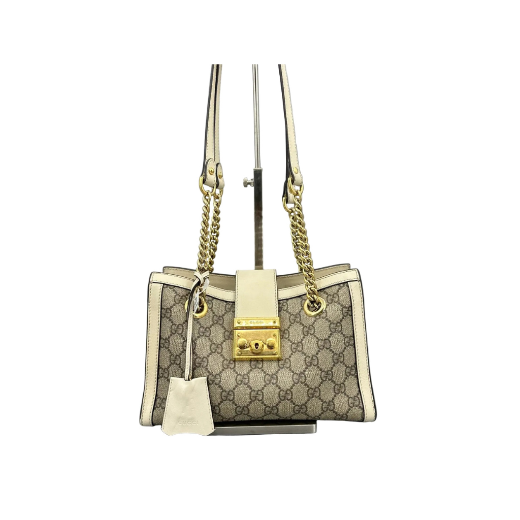Front view of a Gucci Padlock Medium Shoulder Bag in GG Supreme canvas with white leather trim and gold hardware on a display stand.