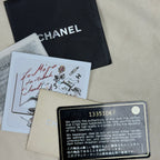 Chanel authenticity card, dust bag, and papers laid out on a beige fabric surface. The black Chanel dust bag is visible at the top, with an authenticity card and a metal plate below it.