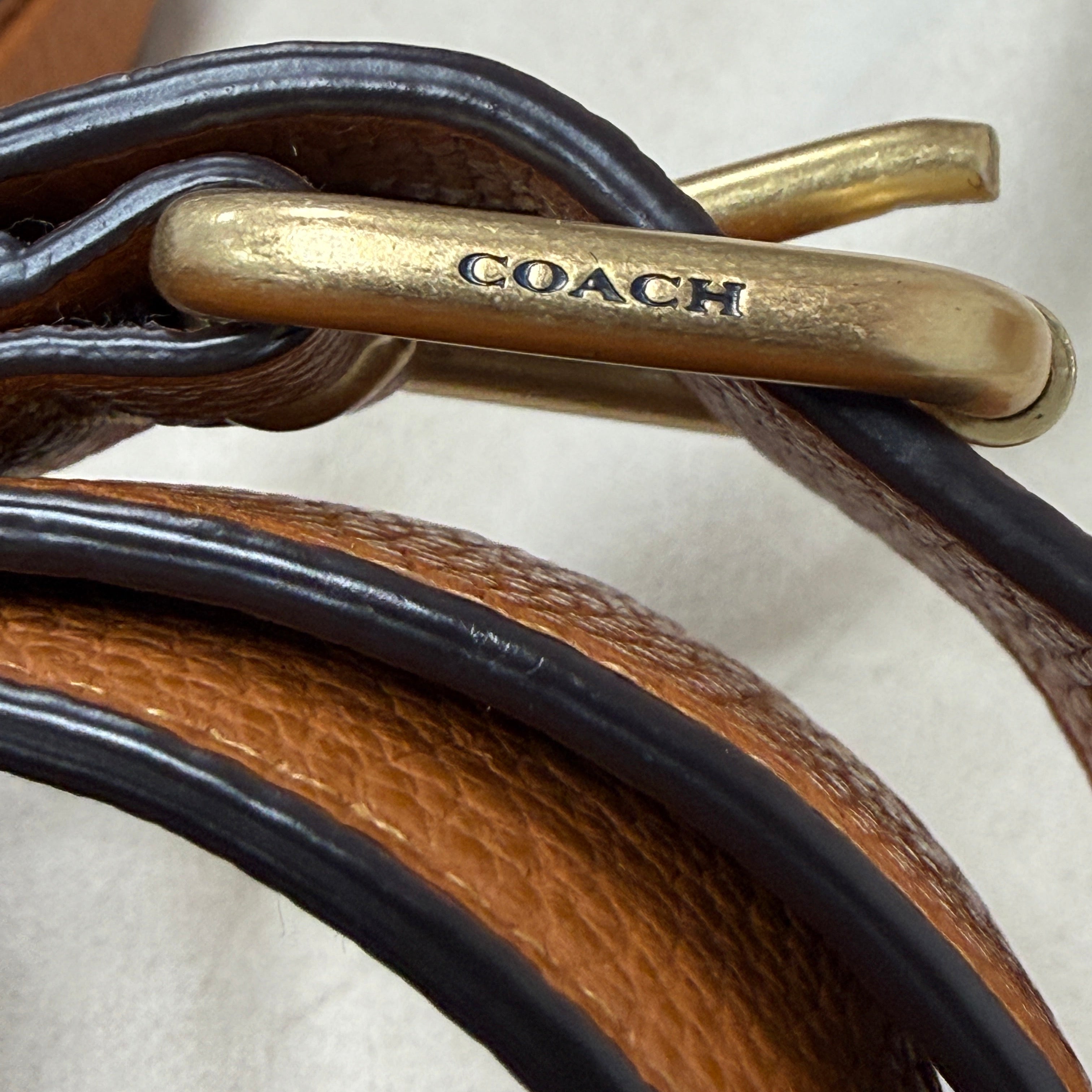 Close-up of a Coach leather strap with a gold-tone metal buckle engraved with the Coach logo, showing brown textured leather and black trim on a light neutral background.