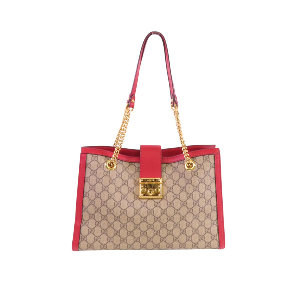 A Gucci GG Supreme tote bag in beige and brown monogram canvas with bright red leather trim and handles, featuring gold-tone hardware and a front lock closure, shown on a plain white background.