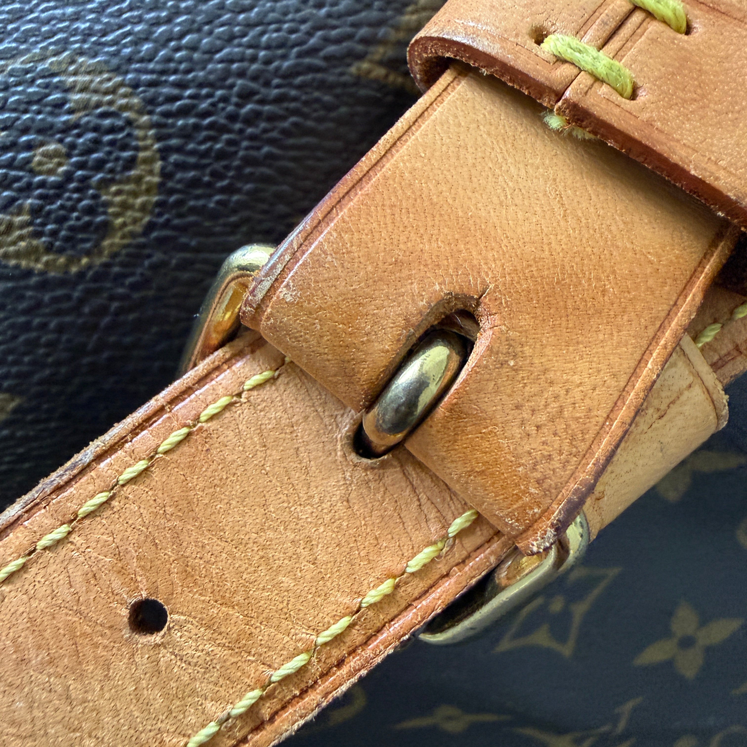 Close-up of a Louis Vuitton monogram canvas bag strap with a tan leather buckle strap, light yellow stitching, and brass hardware.