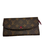 A Louis Vuitton monogram wallet in brown coated canvas with tan LV logos and floral motifs, featuring a small red snap button on the front flap and a red fabric pull tab on the right side, photographed on a plain light background.