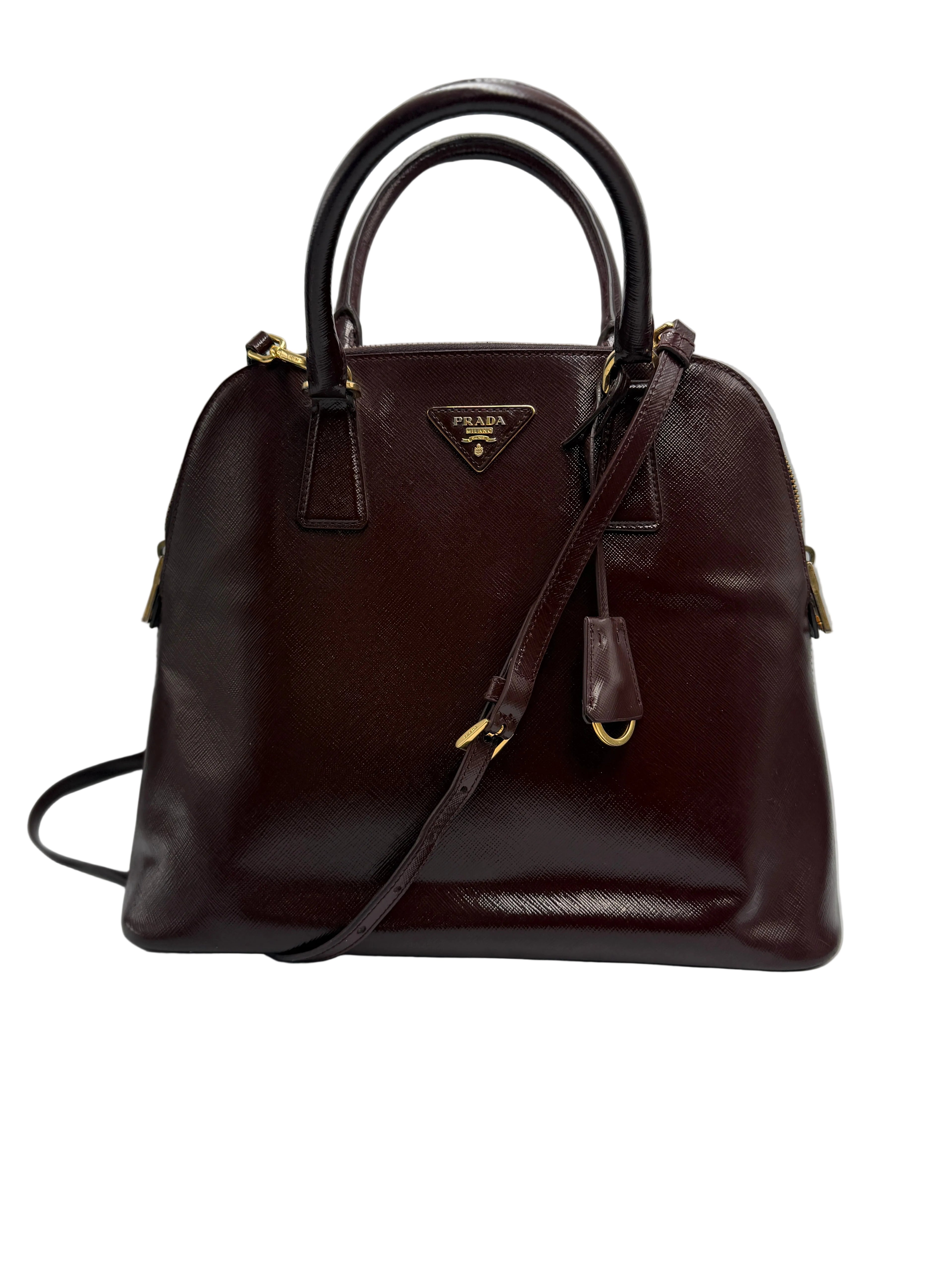 Front view of a Prada Promenade dome satchel in deep burgundy Saffiano leather with top handles, crossbody strap, and gold triangle logo.