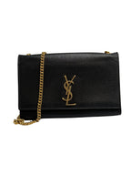 A black pebbled leather Saint Laurent shoulder bag with a gold-tone YSL logo on the front flap and a gold chain strap, photographed against a neutral light gray background.